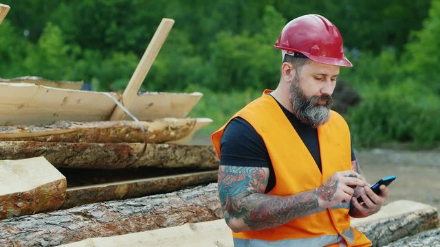  middle-aged man with tattoos and beard enjoys a smartphone, sms writing. He is very serious or upset, sitting on a pile of logs construction