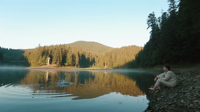 Handsome young thoughtful man in embroidery traditional ukrainian clothes throwing stones into the water and looking on the picturesque mountain lake Synevir. Ukrainian mythology concept. Side view