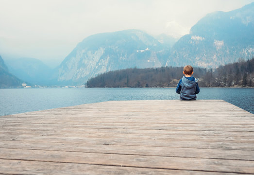 Little boy sits on the wooden pier near the blue mountain lake