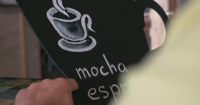Close-up shot of a man writing coffee types on the small chalk board with a drawn cup. Making coffee list for a cafe