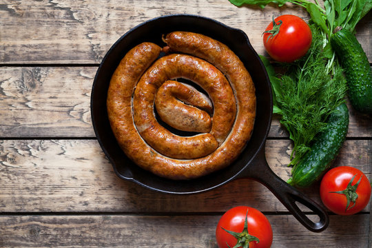 Grilled meat sausage in cast iron pan homemade traditional food with tomatoes, cucumbers and herbs on vintage wooden table background