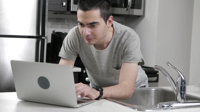 Young man in a home kitchen using a laptop computer on the counter.