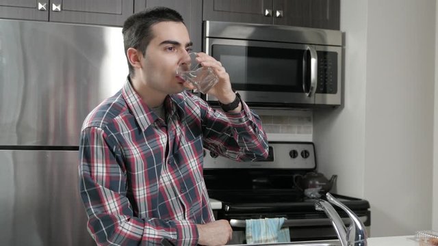 Young man in a home kitchen drinking a glass of water.