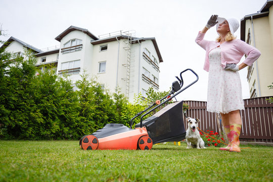 Old Senior Woman Gardener 65 Years Old In Hat, Mowing Grass With An Electric Mower In Garden, Summer Morning