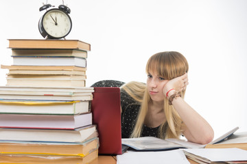 Sad girl student sitting at a desk with a large stack of books and looking at the laptop