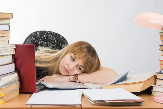 Girl student laid her head on her hand crammed with books and papers lying on the desktop