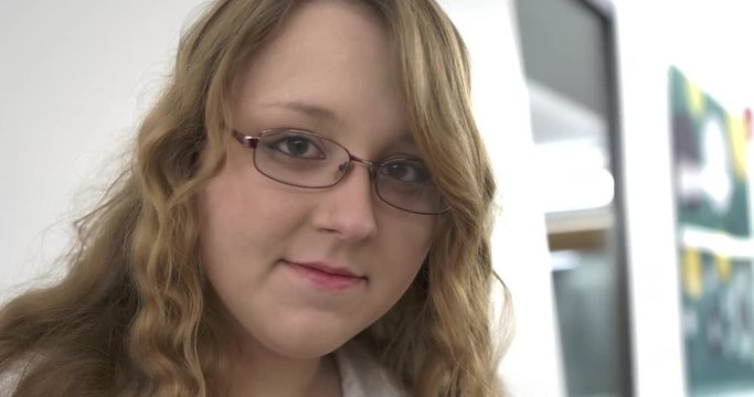 Female with long hair and glasses looking down at phone or book looks up, smiles slightly at the camera, then looks back down.
