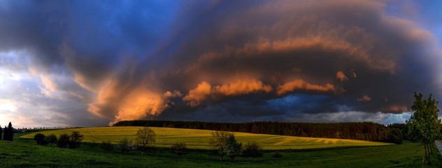 dramatically evolving the cloudy formation lit by the setting sun over the meadow and a field with blooming rape, Zabrehs highlands, North Moravia,Czech republic, panoramic view compiled from six shot