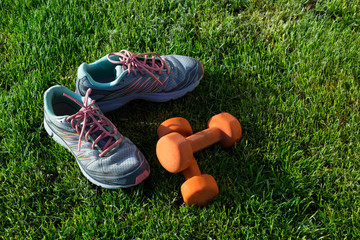 Sports shoes sneakers and pair of orange dumbbells on grass. 