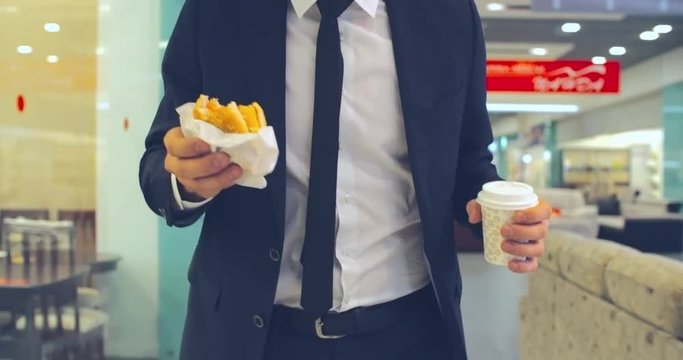 Medium close up of unrecognizable businessman walking in the shopping mall, eating burger and drinking coffee from disposal cup. Suitable for lifestyle, urban workday, food on the go