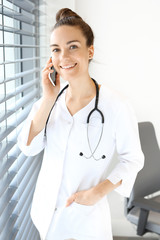 Rozmowa telefoniczna z lekarzem. Lekarka ubrana w biały kitel rozmawia przez telefon stojąc przy oknie.