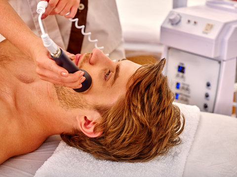 Young man luxuriating on electroporation  facial massage therapy at beauty salon. Man takes care of his face through facing massage.