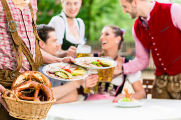 Kellner serviert Essen in bayerischem Biergarten, Freunde essen und trinken im Hintergrund