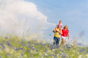 Familie macht Spaziergang auf Wiese mit Blumen, der Vater trägt seine Tochter auf den Schultern