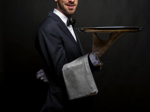 Waiter in black suit holding tray over black background.