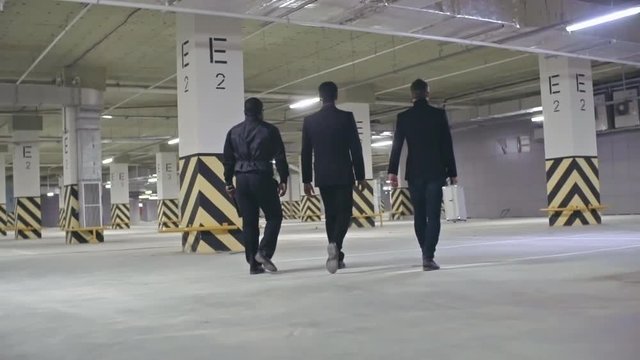 Rear view of three bandits walking in the underground parking; one of them holding metal briefcase and another man carrying baseball bat