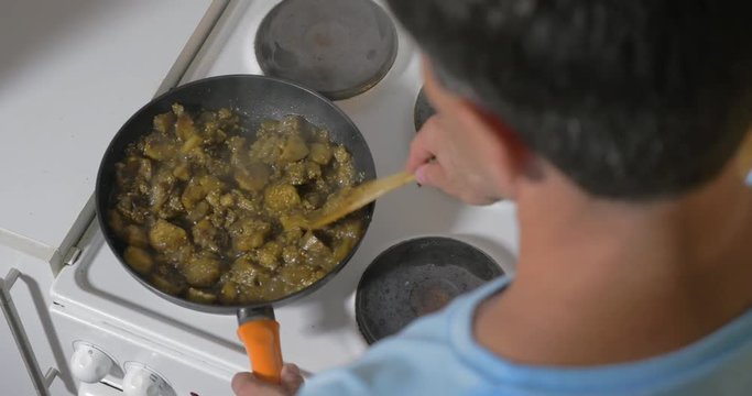 High angle shot of a man cooking vegetarian dish at home. He stirring up egg-plants in the pan and adding cut tomatoes