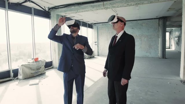 Two businessmen in oculus rift goggles talking and watching plan of future building