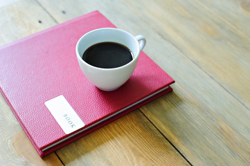 White Cup with coffee standing on the book with the red cover, wooden background.