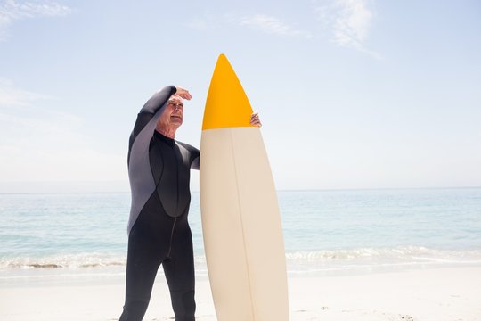 Senior man with surfboard shielding eyes at beach