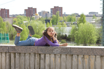 Cute little girl dressed in jeans lying on a high stone parapet and dreaming with closed eyes on the city background
