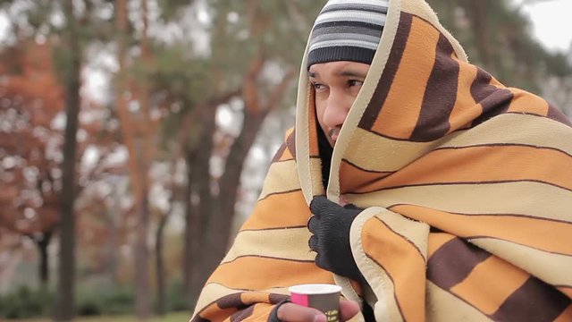 Poor man with sad face hiding from cold in shabby blanket, warming with hot tea