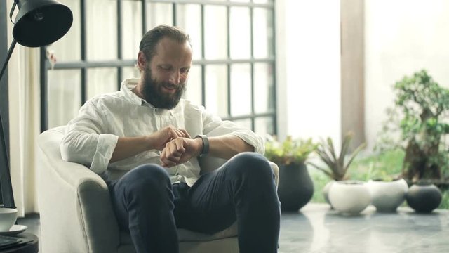 Young, happy man using smartwatch sitting on armchair in living room at home
