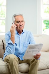 Senior man reading document while talking on phone at home