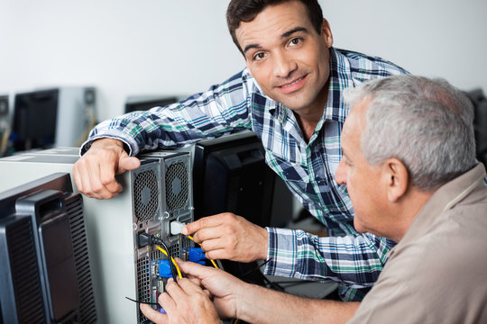 Male Teacher Assisting Senior Man In Fixing Computer