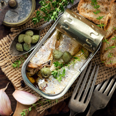 Open tin of sardines in olive oil with capers and thyme on dark rustic wooden background