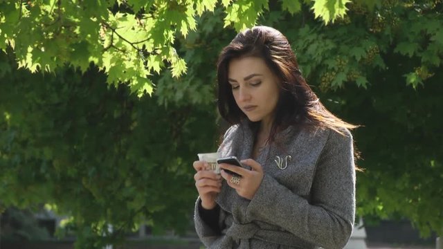 SheThe girl in a park drinking coffee and looking to the smartphone. Then he distracted, as if something she saw, smiled and went back to the phone hands the phone coffee