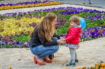 Young lovely mother and her daughter, the girl child, play in the playground on a gravel track among klymb blooming flowers, sunny day with a balloon in her hands, happy family