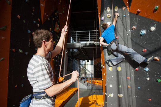 Guy holding a rope with special equipment insuring the climber on rock wall indoors, using belay device and rope