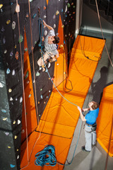 One guy climbing on an indoor rock-climbing wall, the second standing on the ground insuring the climber