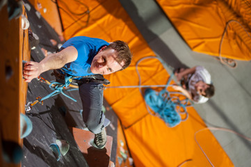 Strong happy male climber practicing climbing on rock wall indoors, view from above. Man standing on the ground insuring the climber
