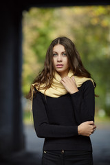 Young romantic brunette woman with long wavy hair posing outdoors in black sweater and yellow scarf with blurry park background