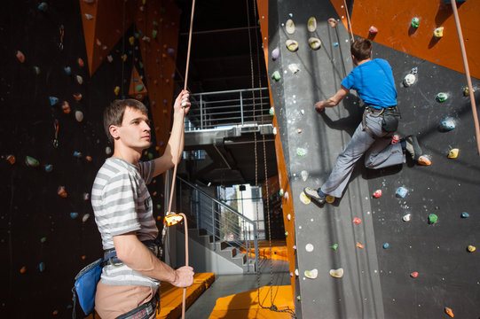 Guy holding a rope with belay device and carbine and belaying the climber on rock wall indoors