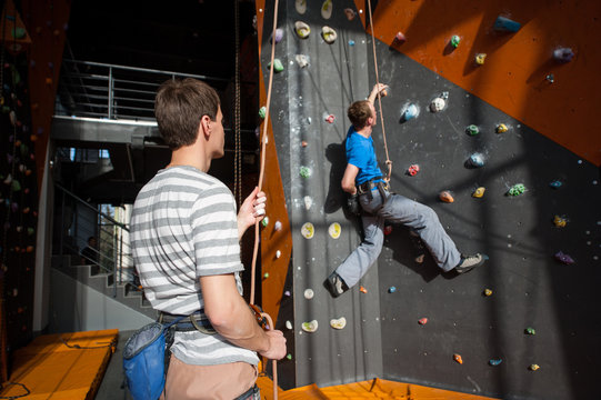 Belayer stands on the ground near rock wall indoors, belaying the climber