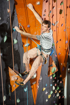 Athletic young male climber is climbing on practical wall indoor, looking to the camera