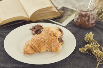 A fresh croissant over plate with chocolate jar to eat while you read a book in a cozy morning