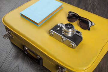 Sunglasses, vintage camera and book on yellow valise on dark background.Travel concept. 