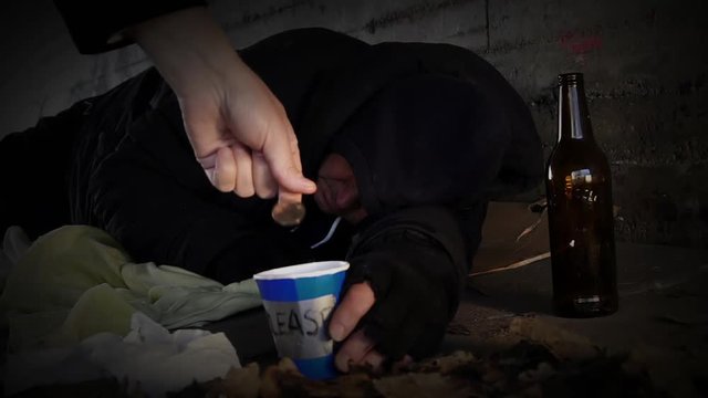 A homeless beggar holding a paper mug and sleeping in a tunnel gets money from a man passing by. Slow motion clip.