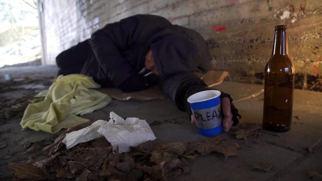 A homeless beggar holding a paper mug and sleeping in a tunnel. Panning shot.