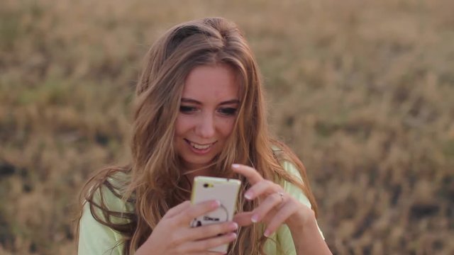 Happy smiling young attractive teenage girl with long blonde hair using smartphone and laughing. Young woman taking pictures in the field at sunset. 