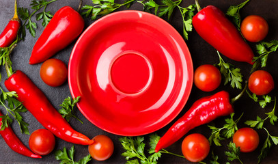 Fresh chili peppers, herbs around red plate on black background