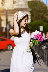 Outdoor portrait of young beautiful happy smiling lady walking on the street. Model wearing stylish white clothes & accessories. Girl looking down & aside.. City lifestyle. Sunny day. Waist up.