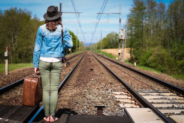 Une jeune femme brune avec un chapeau attend un train au milieu des rails avec une valise à la main