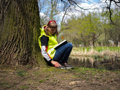Teen girl in the park with a book. Concept - exam preparation, difficulty learning, school. The girl has her hair dyed red. Bright jacket. Teen girl in the park with a book