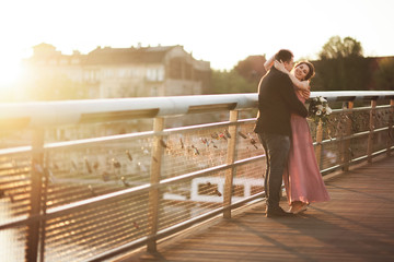 Stylish loving wedding couple, groom, bride with pink dress kissing and hugging on a bridge at sunset
