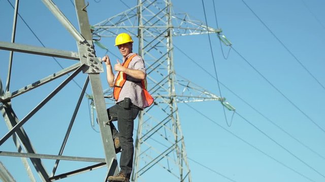 Technician in a safety vest and hard hat standing on the side of a high tension electrical tower visually inspects the situation and give the viewer a thumbs up gesture.